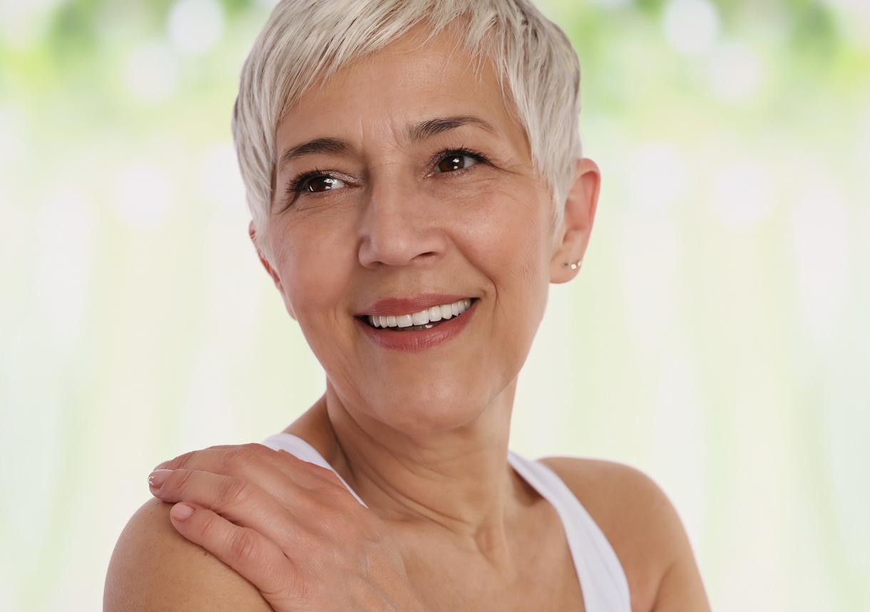 Mature woman with short gray hair wears a white tank top as she shyly shows off her perfect smile