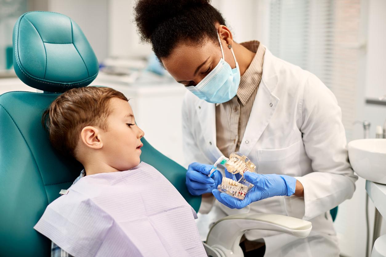 Cute little boy sits in dentist chair as he watches a black female dentist show him how to brush properly using an artificial set of teeth.
