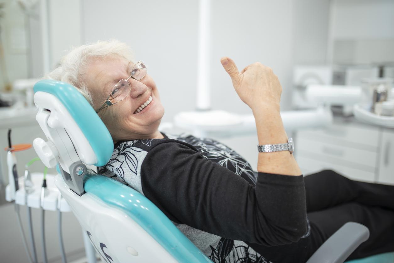 Senior dental patient relaxes in cushioned dental chair and gives a thumbs up as she smiles