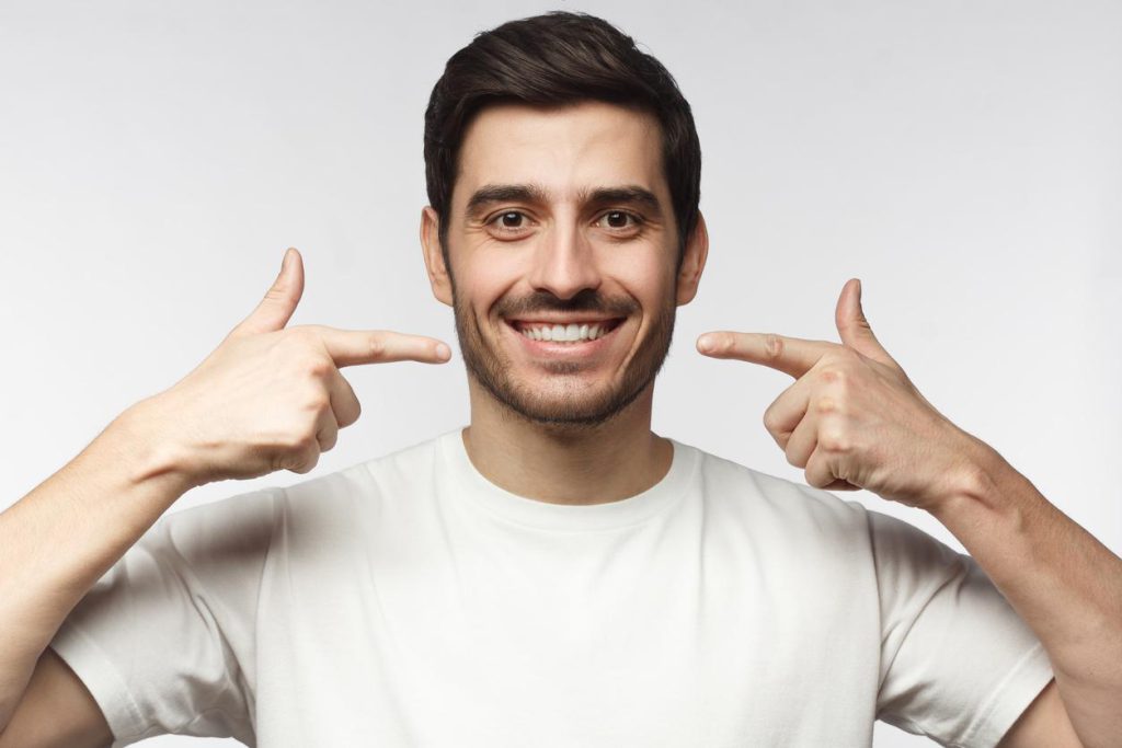 Dark-haired man in white t-shirt used both hands to point at his own beautiful smile - Cosmetic Dentist in Lakeview