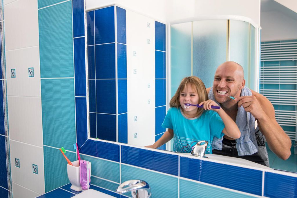 A dad teaching his young daughter how to brush her teeth.
