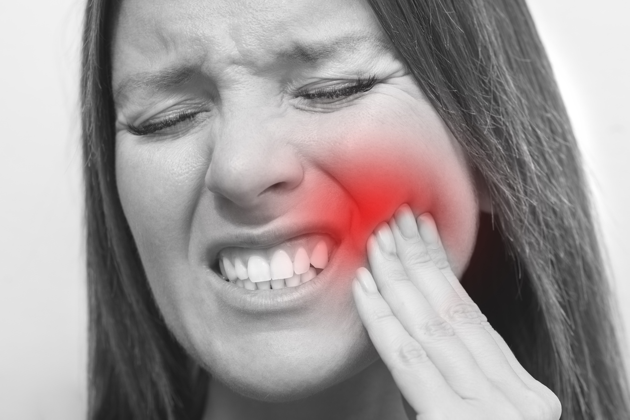 A black and white image of a young woman suffering severe dental pain. She is grimacing and holding the tips of the fingers on her left hand to her left jaw. 