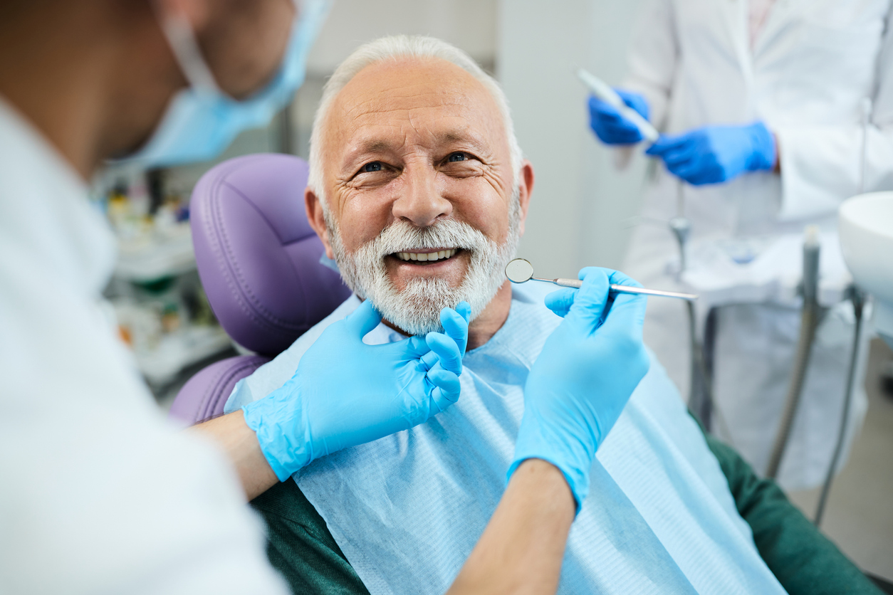 An older white male with gray hair and goatee smiling broadly at the dentist or hygienist performing a dental checkup.