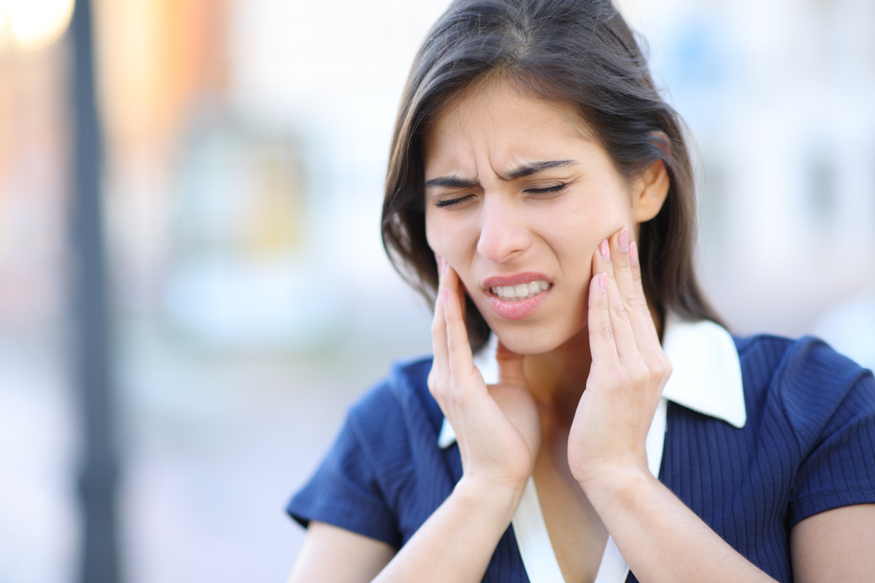 Image of a young, dark-haired woman grimacing in pain with both hands on her jaws, probably suffering from TMJ.