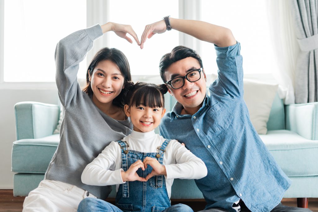 An Asian-American wife, husband, and small daughter sitting on the floor of their home in front of a couch. The smily parents have formed a heart shape with their arms, and the smiling young daughter is doing the same thing with her hands and fingers.